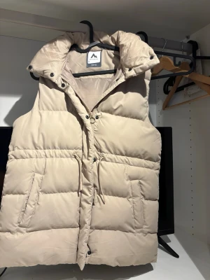 Beige dunväst med huva från McKinley - Snygg beige dunväst från McKinley med dragkedja, tryckknappar och justerbar dragsko i midjan. Västen har en hög krage och en vadderad huva för extra värme. Perfekt för lager-på-lager och chill höst- och vinterstil. Jag är i xs och så sitter den på mig, passar säkert större.