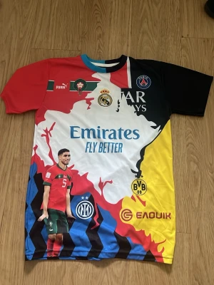 Flerfärgad fotbollströja Hakimi #2 - Unik fotbollströja med Hakimi #2, flera klubbmärken som PSG, Real Madrid, Inter, Dortmund och Marockos landslag. Flerfärgad design i rött, svart, gult, blått och vitt. Tillverkad i lätt och ventilerande polyester, perfekt för fotbollsträning eller som supportertröja. Vill du sticka ut på sommaren så har du hittat rätt tröja🤝