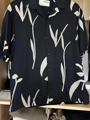 Blå skjorta med vita blad Selected Homme - Snygg blå  skjorta från Selected Homme med vita bladliknande mönster. Skjortan har korta ärmar, klassisk krage och knäppning framtill. Perfekt för en avslappnad och trendig look. Tillverkad i ett mjukt och lätt material.