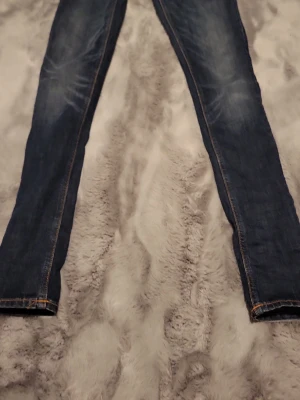 Mörkblå skinny jeans från Nudie - Snygga mörkblå skinny jeans från Nudie med klassiska orange sömmar och vågig brodyr på bakfickan. Jeansen har en smal passform och är tillverkade i denim med lätt slitning på låren. Perfekta för en trendig och avslappnad look.
