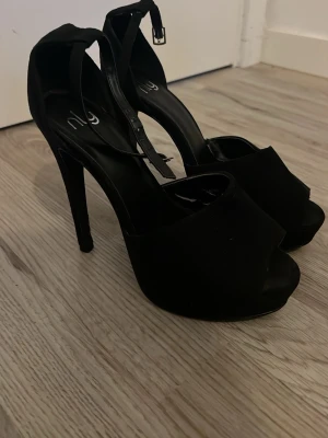 Svarta höga pumps från NLY - Säljer ett par svarta pumps från NLY med öppen tå och hög klack. Skorna har en bred rem över foten och justerbart ankelband med spänne. Snygg och stilren design som passar till många olika outfits.