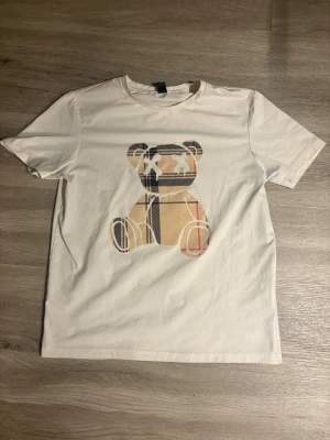 Vit t-shirt med rutigt nalletryck - Vit t-shirt med ett stort tryck av en nallebjörn i beige, svart och rött rutmönster på bröstet. Klassisk passform och rund halsringning. Perfekt för dig som gillar lekfulla och trendiga motiv.