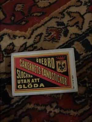 Örebro säkerhetständstickor ask - Retro tändsticksask med klassisk Örebro-etikett i svart, rött och gult. Framsidan har text och lejonemblem, baksidan listar gamla etikettserier. Perfekt för samlare eller som dekoration.