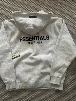 Essentials hoodie - Snygg ljusgrå hoodie från Essentials Fear of God med stor svart logga på ryggen. Klassisk passform med huva och ribbade muddar. Tillverkad i mjukt bomullsmaterial som känns skönt mot huden. Perfekt för en avslappnad streetwear-look.