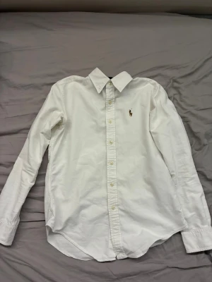 Vit skjorta från Ralph Lauren - Klassisk vit skjorta från Ralph Lauren med broderad logga på bröstet. Skjortan har lång ärm, knappar framtill och är tillverkad i 100% bomull. Perfekt till både jeans och chinos för en clean och tidlös look. Priset kan diskuteras.