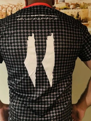Svart sporttröja Club Deportivo Palestino / M - Svart sporttröja med hexagonmönster, röda och gröna ränder samt klubbens emblem på bröstet. Tryck i vitt på ryggen och ärmen, samt text på arabiska och spanska. Perfekt för fotboll eller träning.