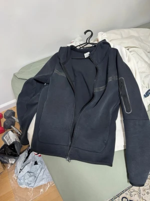Svart hoodie med dragkedja från Nike - Snygg svart hoodie från Nike med trasig dragkedja. Hoodien har diskreta detaljer på ärmen och bröstet samt en ficka med dragkedja på ena ärmen. Perfekt för en sportig och avslappnad stil.
