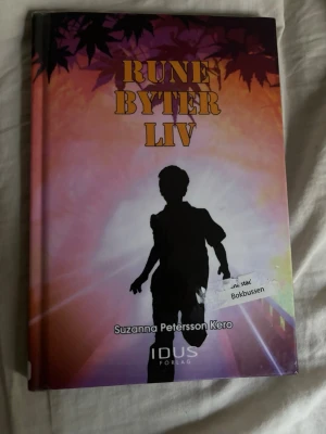 Rune byter liv av Suzanna Petersson Kero - En färgstark inbunden bok med titeln 'Rune byter liv' av Suzanna Petersson Kero. Omslaget har en siluett av ett barn mot en bakgrund i lila, orange och rosa toner med lövdetaljer. Boken handlar om Rune som är adopterad och söker sin plats i tillvaron.