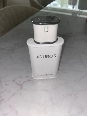 Kouros parfym från Yves Saint Laurent - Kouros parfym från Yves Saint Laurent i en stilren vit flaska med silverfärgad topp. Flaskan har en modern, fyrkantig form och är tillverkad i plast och metall. Perfekt för dig som gillar klassiska dofter med lyxig känsla.