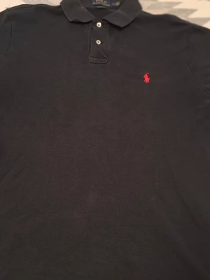 Svart pikétröja från Ralph Lauren - Klassisk svart pikétröja från Ralph Lauren med röd broderad logga på bröstet. Tröjan har krage, två knappar framtill och är tillverkad i bomull. Perfekt för en stilren och avslappnad look.
