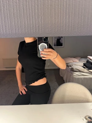 Svart ribbad croppad topp - Svart ribbad croppad topp med korta ärmar och vågig kant nertill och vid ärmslut. Enkel och stilren design som passar perfekt till jeans eller kjol. Mjuk och stretchig för en bekväm passform.