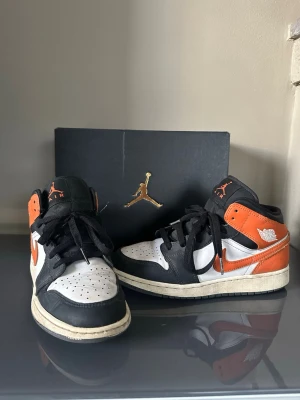 Air Jordan 1 Mid svart orange vit Stl 38 - Snygga Air Jordan 1 Mid sneakers i svart, orange och vitt. Klassisk siluett med svart swoosh, orange detaljer och vita paneler. Skorna har svarta skosnören och den ikoniska Air Jordan-loggan på sidan. Perfekta för dig som gillar streetwear och vill sticka ut.
