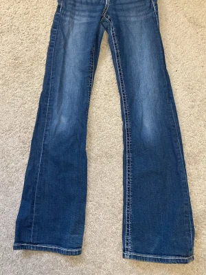 Blå bootcut jeans med slitningar - Ett par klassiska blå bootcut jeans i denim med lätt slitna detaljer framtill. Jeansen har normal midja och vida ben från knät och ner. Perfekta för en avslappnad och trendig look.