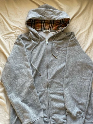 Grå zip hoodie  - Snygg grå zip hoodie från Burberry med klassiskt rutigt foder i huvan och broderad logga på bröstet. Tillverkad i mjuk bomullsblandning, har dragsko i huvan och fickor framtill. Perfekt för en chill och stilren look.