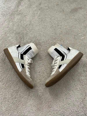 Maison Margiela Sock Gat Sneakers - Unika vita sneakers med ribbad textil och svarta ränder, beige mockadetaljer och klassisk brun gummisula. Skorna har en strumpliknande design och svart kant runt öppningen. Perfekta för dig som vill sticka ut med din stil.