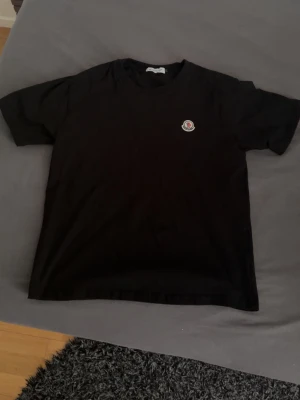 Svart Moncler t-shirt med logga - Snygg svart t-shirt från Moncler med klassisk rund hals och korta ärmar. På bröstet sitter Monclers ikoniska broderade logga i vitt, rött och blått. Perfekt basic-plagg med exklusiv känsla och stilren design.