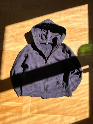 Mörkblå hoodie från Polo Ralph Lauren - Mysig mörkblå hoodie från Polo Ralph Lauren med dragkedja framtill och klassisk huva. Liten röd logga broderad på bröstet och två fickor fram. Tillverkad i mjuk fleece som känns skön mot huden. Perfekt för en avslappnad och stilren look.
