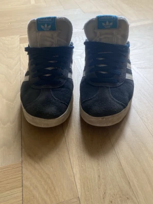 Adidas Gazelle blåa sneakers - Klassiska Adidas Gazelle sneakers i blå mocka med vita detaljer och blå logga på plösen. Skorna har de ikoniska tre vita ränderna på sidan och vit sula. Perfekta för en stilren och sportig look.