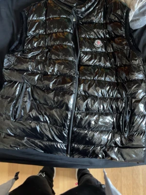 Svart glansig dunväst från Moncler - Säljer en svart, glansig dunväst från Moncler med klassisk logga på bröstet. Västen har två dragkedjefickor och quiltad design. Perfekt för lager-på-lager och riktigt snygg till streetwear-stilen.