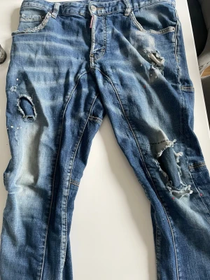 Blå slitna jeans med hål och stänk - Säljer ett par blå DSQUARED2 jeans med slitningar, hål och färgstänk för en cool och trendig look. Jeansen har klassisk femficksmodell och raka ben. Perfekta för dig som gillar streetstyle och vill sticka ut lite extra.
