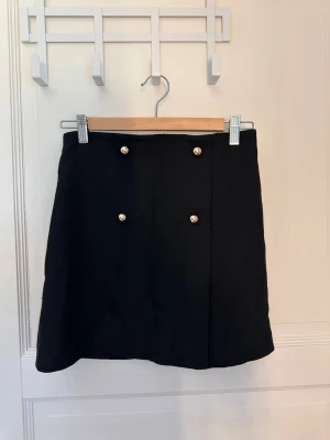 Svart kortkjol med guldknappar - Stilren svart kortkjol med fyra dekorativa guldknappar framtill. Kjolen har en rak passform och är perfekt för dig som gillar en klassisk men trendig look. Passar snyggt till både stickad tröja och t-shirt. Materialet känns stabilt och håller formen bra.