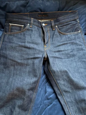 Mörkblå jeans från Nudie Jeans - Snygga mörkblå jeans från Nudie Jeans i klassisk rak modell. Jeansen har kontrastsömmar i orange och fem fickor, inklusive myntficka. Tillverkade i kraftig denim av bomull, perfekta för en stilren och tidlös look.
