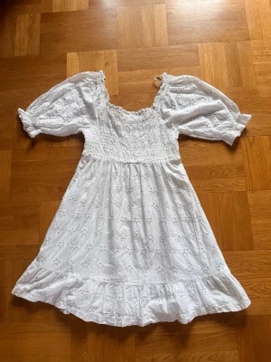 Vit kort klänning med broderade blommor - Söt vit kortklänning med broderade blommor och volangkant. Klänningen har puffiga kortärmar och smockad överdel som ger en fin passform. Perfekt för varma dagar och skolavslutning 