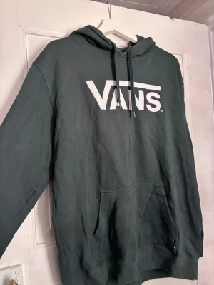 Mörkgrön hoodie från Vans med logga - Snygg mörkgrön hoodie från Vans med stor vit logga på bröstet. Klassisk modell med huva och dragsnören samt en stor magficka framtill. Perfekt för en avslappnad streetstyle-look. Tillverkad i mjuk bomullsblandning för extra komfort.