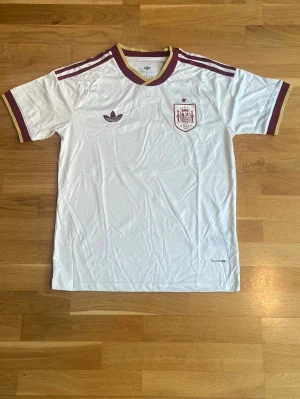 Vit Spanien fotbollströja Adidas - Snygg vit fotbollströja från Adidas med Spaniens landslagsemblem på bröstet. Tröjan har vinröda och guldiga detaljer vid krage, ärmslut och axlar. Klassisk Adidas-logga och broderat märke. Perfekt för fotbollsfans!