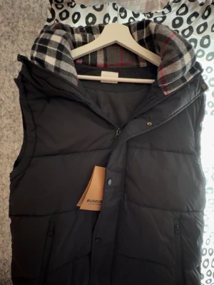 Svart dunväst från Burberry - Stilren svart dunväst från Burberry med klassiskt rutigt foder i huvan. Västen har dragkedja och tryckknappar framtill samt två fickor med dragkedja. Perfekt för lager på lager och ger en lyxig känsla till din outfit.