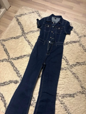Mörkblå denim jumpsuit från SHEIN - Snygg mörkblå denim jumpsuit från SHEIN med korta ärmar, krage och knappar framtill. Har fickor på bröstet och markerad midja. Tillverkad i stretchigt jeanstyg som ger en bekväm passform. Perfekt för dig som vill ha en cool och trendig look.
