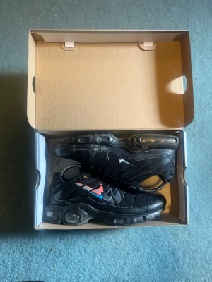Nike Air Max Plus svarta sneakers - Svarta Nike Air Max Plus sneakers med mesh och syntetmaterial, ikoniska vågformade detaljer och synlig Air Max-dämpning i sulan. Swoosh-loggan är blå och röd på sidan, vilket ger en unik touch. Skorna har snörning och en sportig, chunky siluett.skorna är välanvända men inga hål,pris kan diskuteras 