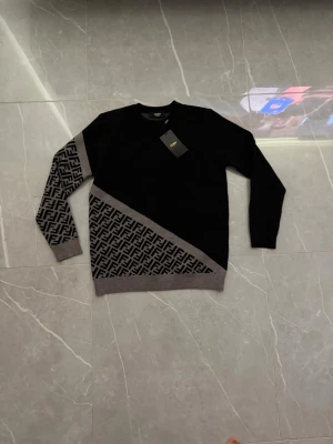 Svart Fendi tröja med FF-mönster - Svart tröja från Fendi med gråa ribbade muddar och diagonal FF-logomönster på ena ärmen och nedre delen. Rund hals och klassisk passform. Materialet ser ut att vara stickat och ger en lyxig känsla. Perfekt för dig som gillar exklusiv streetstyle.