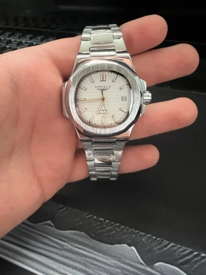 Klockan  - Stilren klocka från Longlux i silverfärgat rostfritt stål med automatisk urverk. Urtavlan är vit med datumvisning och tydliga markeringar. Länken är robust och klockan har en transparent baksida som visar mekaniken. Perfekt accessoar för en modern look.