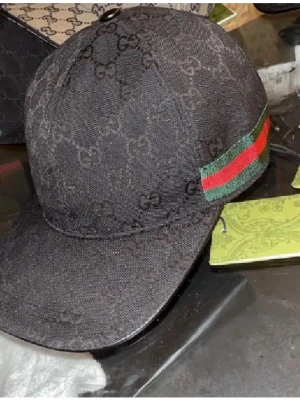 Svart Gucci keps med GG-mönster - Svart keps från Gucci med klassiskt GG-monogram över hela kepsen. På sidan finns en grön och röd randig detalj som ger en lyxig känsla. Kepsen har böjd skärm och är tillverkad i textilmaterial.