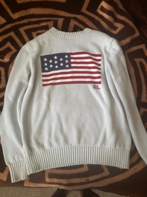 Ljusgrå stickad tröja Ralph Lauren - Snygg ljusgrå stickad tröja från Ralph Lauren med amerikansk flagga och RL-broderi på bröstet. Tröjan har rund halsringning, långa ärmar och ribbstickade muddar. Perfekt för dig som gillar klassisk och stilren design.