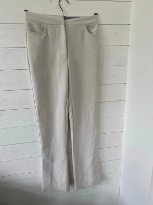 Beiga kostymbyxor med slits - Stilrena beiga kostymbyxor med raka ben och diskret slits nertill. Byxorna har hög midja. Perfekta att matcha med en skjorta eller stickad tröja. Materialet känns mjukt och följsamt.