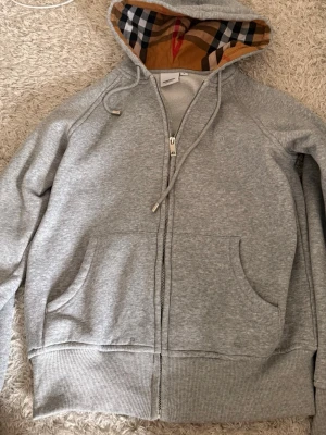 Grå zip-hoodie från Burberry 160cm - Säljer en grå zip-hoodie från Burberry med klassiskt rutigt foder i huvan och broderad logga på bröstet. Hoodien har dragsko i huvan, två fickor fram och ribbade muddar. Perfekt för en chill och stilren look.