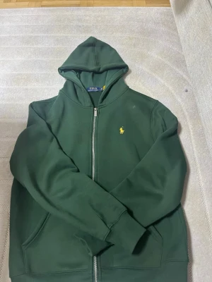 Grön hoodie från Polo Ralph Lauren - Mörkgrön hoodie med dragkedja från Polo Ralph Lauren. Klassisk gul logga broderad på bröstet, huva och två fickor framtill. Tillverkad i mjukt sweatshirtmaterial, perfekt för en chill och avslappnad stil.