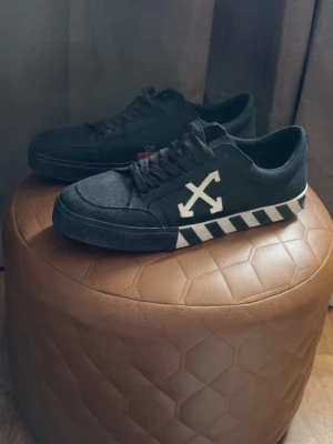 Off-White svarta sneakers med pilar - Svarta sneakers från Off-White med vita pilar på sidan och svartvita ränder på sulan. Skorna har lågt skaft, snörning och är tillverkade i canvas och mocka. En ikonisk streetwear-modell som sticker ut med sin unika design.
