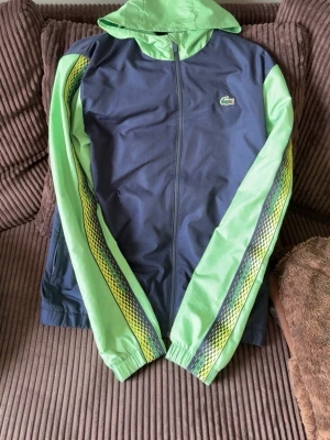 Lacoste vindjacka i blått och grönt - Snygg vindjacka från Lacoste i blått med ljusgröna ärmar och huva. Ärmarna har coola grafiska mönster i gult och svart längs sidorna. Jackan har dragkedja framtill och klassisk Lacoste-logga på bröstet. Perfekt för en sportig och trendig look.