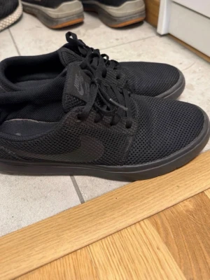 Svarta Nike sneakers mesh - Svarta Nike sneakers med lågt skaft och klassisk swoosh på sidan. Skorna har ovandel i mesh för extra andningsförmåga och en svart sula som matchar resten av skon. Perfekta för en clean och sportig look.