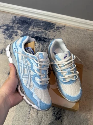 ASICS Gel-1090 vita och blå sneakers - Fräscha ASICS Gel-1090 sneakers i vitt och ljusblått med mesh- och syntetpaneler. Klassisk låg modell med tydliga ASICS-stripes på sidorna, vadderad plös och häl samt vit sula med blå detaljer. Perfekta för en sportig och trendig look.
