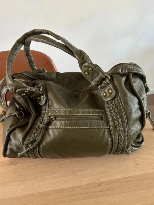 Olivgrön handväska med flätade detaljer - Snygg olivgrön handväska i skinnimitation med flätade detaljer och nitar. Väskan har dubbla handtag, dragkedjeficka framtill och dekorativa sömmar. Perfekt för dig som gillar en cool och trendig look.