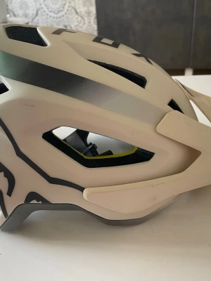 Fox Speedframe MTB-hjälm beige - Snygg benvit Fox Speedframe MTB-hjälm med svart grafik och justerbart visir i tre lägen. Hjälmen har MIPS-system för extra skydd, urtagbara och tvättbara liners samt optimal ventilation genom EPS-kanaler. Passar perfekt för mountainbike och har 360° fastspänningssystem.  Vikt 385 g
