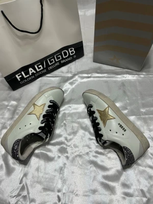 Golden Goose sneakers med glitterdetaljer - Snygga vita Golden Goose sneakers med guldstjärna på sidan, svarta skosnören och glitterdetaljer på hälen. Klassisk låg modell med slitstark gummisula och coola detaljer som gör dem unika. Perfekta för dig som vill sticka ut med din stil.
