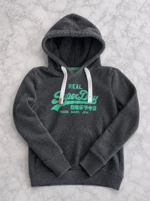 Mörkgrå Superdry hoodie med tryck - Mörkgrå hoodie från Superdry med turkost tryck och japanska tecken på bröstet. Har vit dragsko i huvan och klassisk känguruficka framtill. Mjuk insida och ribbade muddar vid ärmslut och nederkant. Perfekt för en chill och avslappnad stil.
