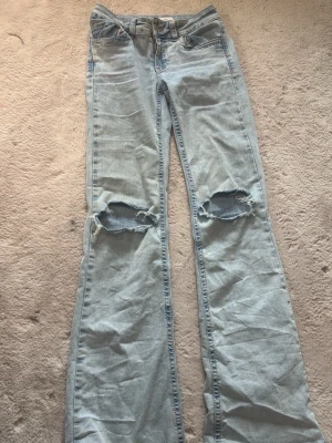 Ljusblå bootcut jeans med hål - Säljer ett par ljusblå bootcut jeans med slitna hål över knäna och broderade detaljer på bakfickorna. Jeansen har klassisk femficksdesign och är tillverkade i denim. Perfekta för en avslappnad och trendig look. Har även sprättat ut dom där nere för dom ska bli längre är 1,73 så dom är perfekt långa dom kostar 700 men säljer dom billigare 