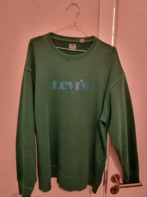 Grön sweatshirt från Levi's - Snygg grön sweatshirt från Levi's med blå logga på bröstet. Tröjan har rund halsringning, långa ärmar och ribbade muddar vid ärmslut och nederkant. Perfekt för en avslappnad och trendig stil.