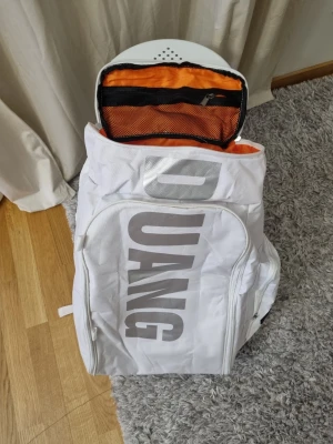 Vit DUANG ryggsäck med orange foder - Snygg och stor vit ryggsäck från DUANG med stort tryck på framsidan och orange innerfoder. Ryggsäcken har flera fack, inklusive ett nätfack med dragkedja på insidan. Perfekt för dig som vill sticka ut med en unik väska.